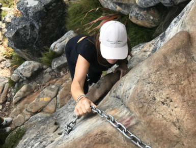 A person climbing a rocky path using a metal chain for support during the India Venster guided hike, organized by Muki Venture. The person is wearing a white cap and a black outfit, with a grassy background and rugged rocks around them