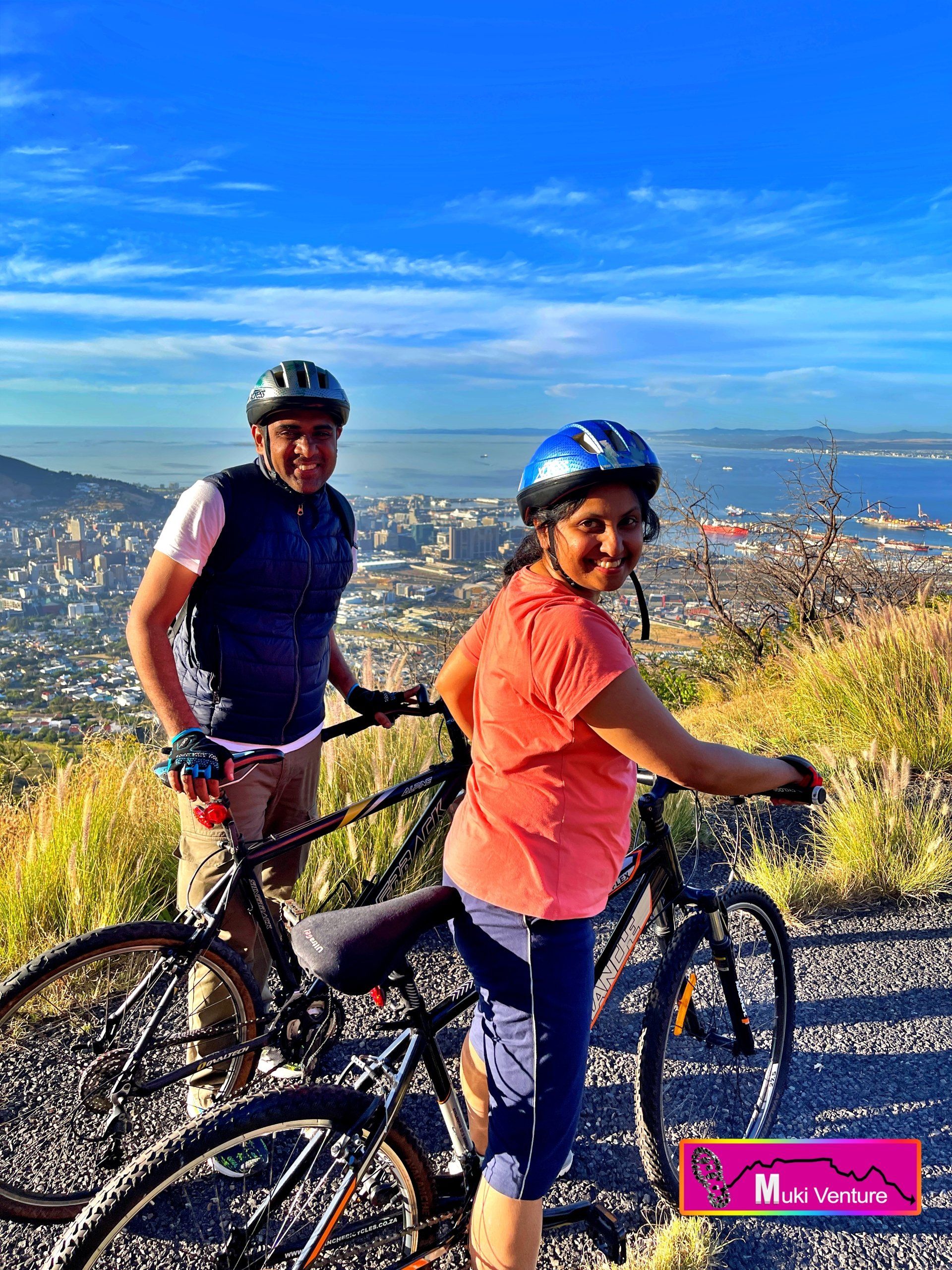 The image shows a couple on a mountain bike tour in a picturesque and mountainous landscape. The couple is standing next to their bikes, both wearing cycling gear and helmets. They appear to be taking a break and enjoying the scenic view, as they stand on a path surrounded by lush green trees and beautiful rolling hills. The couple seems to be in good spirits, with smiles on their faces, suggesting that they are having a great time. The sunlight filters through the tree leaves, creating a warm and inviting atmosphere. The mountains in the background provide a majestic backdrop to the scene, enhancing the feeling of adventure and exploration. Overall, the image portrays a couple embracing the joy and excitement of a mountain bike tour, surrounded by the beauty of nature.