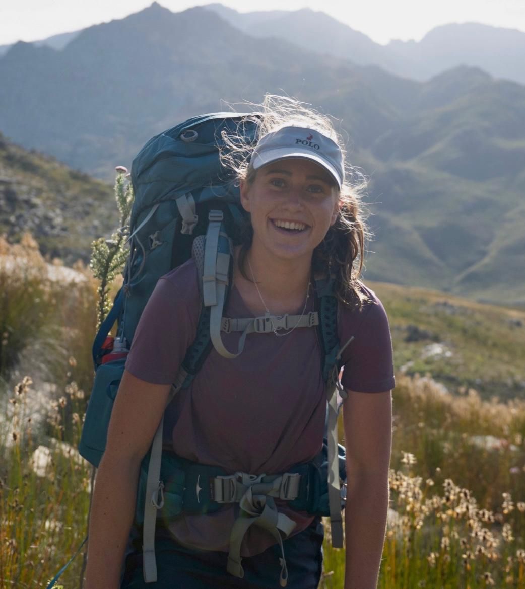 A woman with a backpack is standing in a field with mountains in the background.