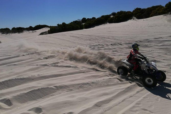 The image shows an off-road vehicle (quad bike) speeding across the Atlantis Sand Dunes, kicking up sand trails behind. These dunes, located near Cape Town, South Africa, are a popular spot for outdoor activities such as sandboarding, quad biking, and hiking.  For a guided hiking experience focused on environmental education, the Atlantis Dunes also offer opportunities to explore the unique flora and fauna of this coastal desert environment. Guided tours here typically include discussions on the region's biodiversity, its conservation efforts, and the ecological significance of sand dune habitats, combining adventure with educational insights into environmental stewardship.