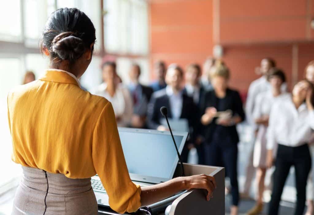 Woman giving presentation at podium, facing audience; laptop, microphone present; indoor setting.