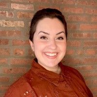 Woman with short dark hair, smiling, wearing a rust-colored shirt, in front of a brick wall.