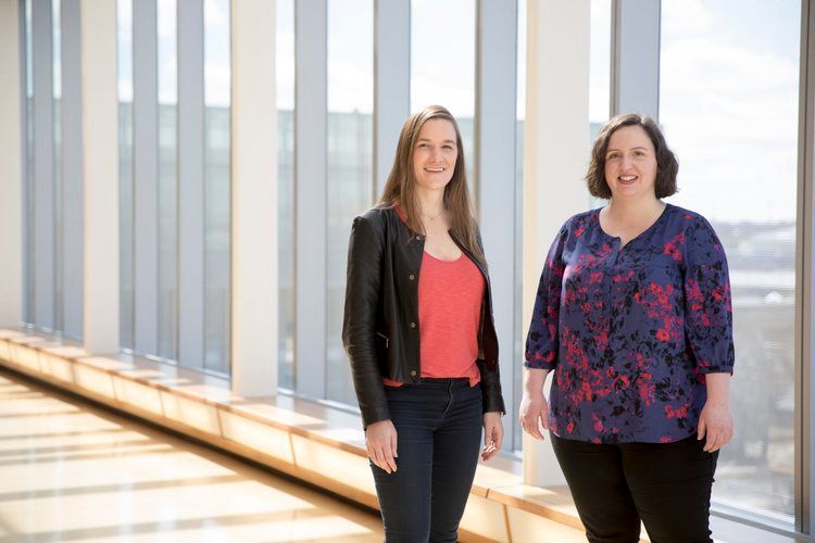 Two women standing in a hallway with large windows, one in a black jacket and jeans, the other in a floral top.
