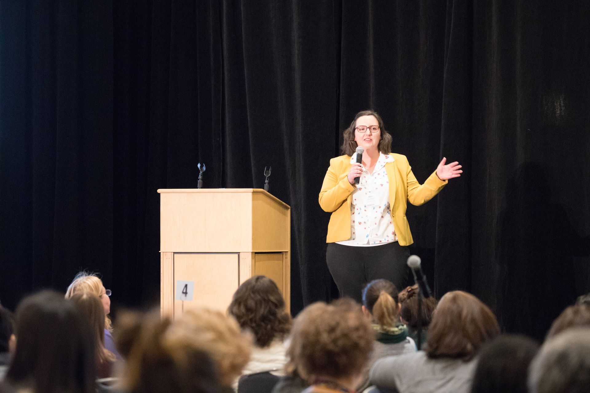 Woman in yellow blazer speaks at a podium to an audience in a darkened auditorium.