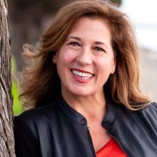 Woman smiles outdoors, leaning against a tree. She wears a black jacket over a red top.