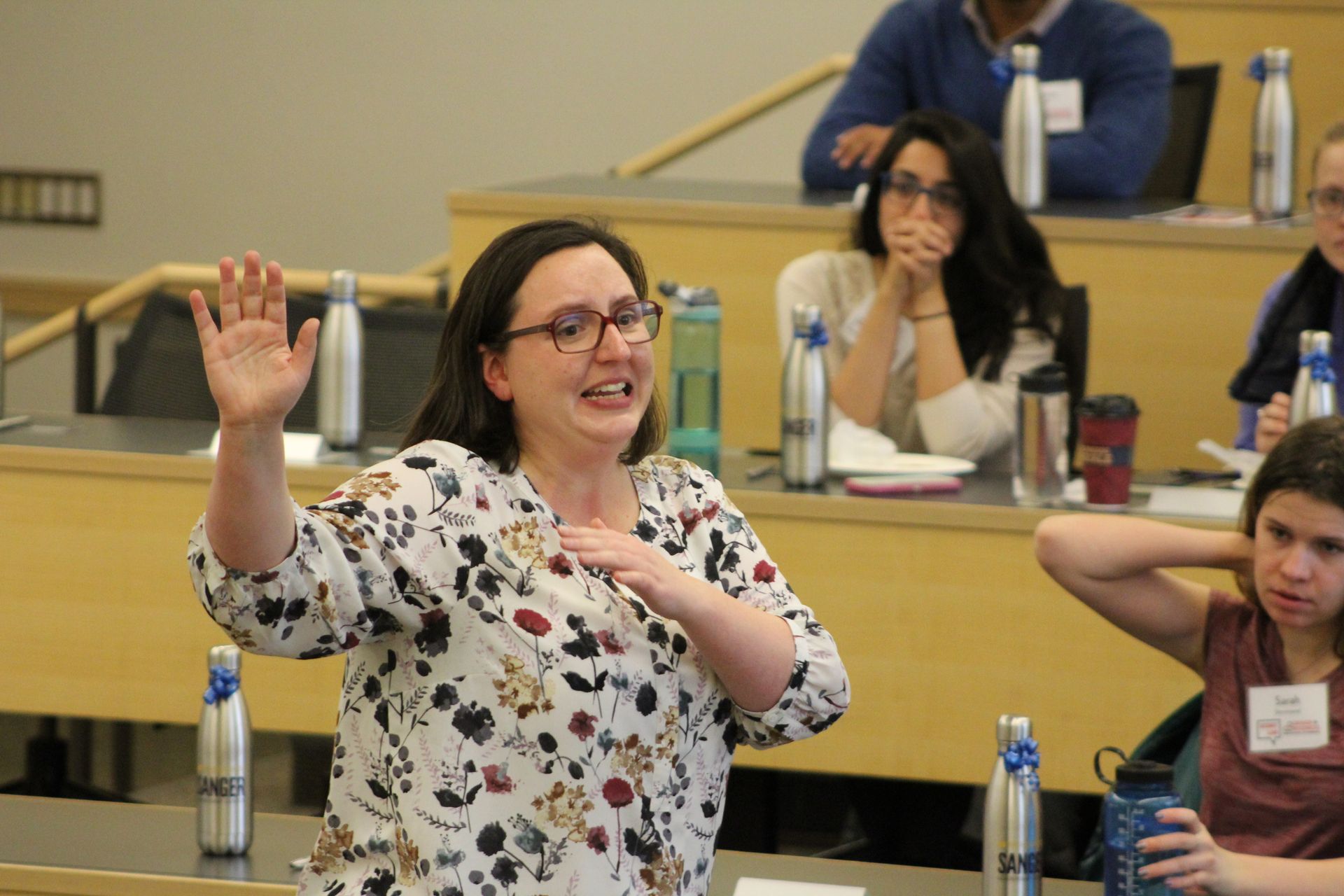 Woman in floral shirt raising hand and speaking in a lecture hall; others watch.