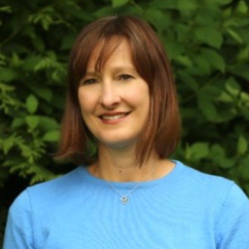 Woman with reddish-brown hair, smiling, wearing a light blue sweater, with a green background.