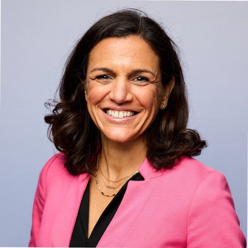 Woman with dark hair smiles, wearing a pink blazer over a black top, with a light blue background.