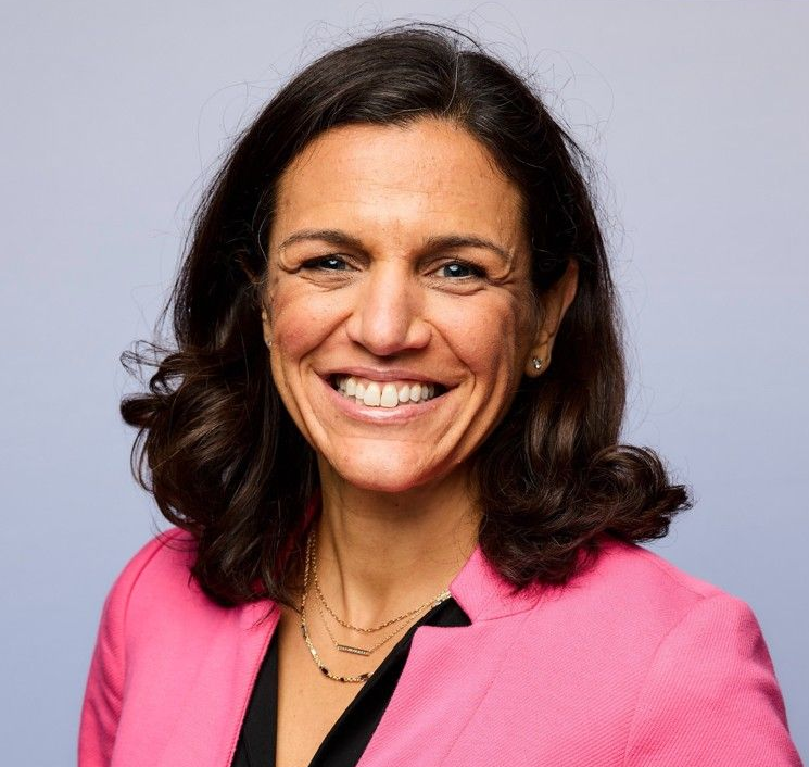 Woman in pink blazer smiles, wearing layered gold necklaces.
