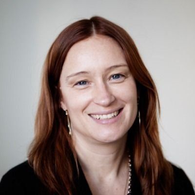 Woman with long auburn hair smiles at the camera, wearing a black shirt and silver earrings, indoors.