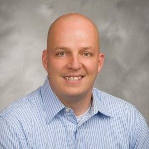 Bald man in a light blue striped shirt smiling at the camera against a grey gradient background.