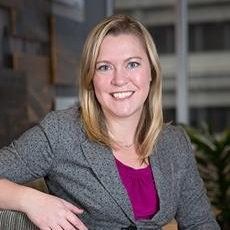 Woman with blonde hair wearing a gray blazer and pink shirt, smiling.