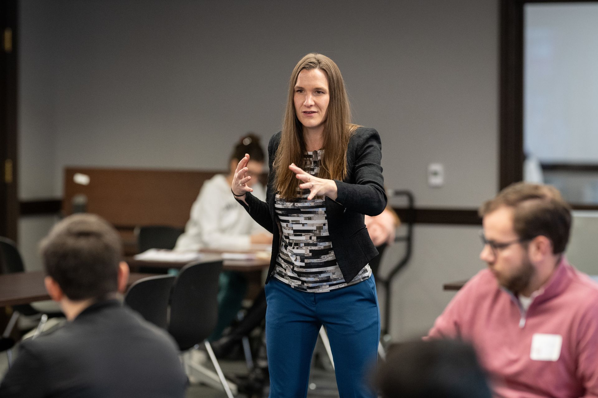 Woman lecturing to a group, gesturing with hands. She's wearing a blazer, patterned shirt, and blue pants. People are seated at tables.