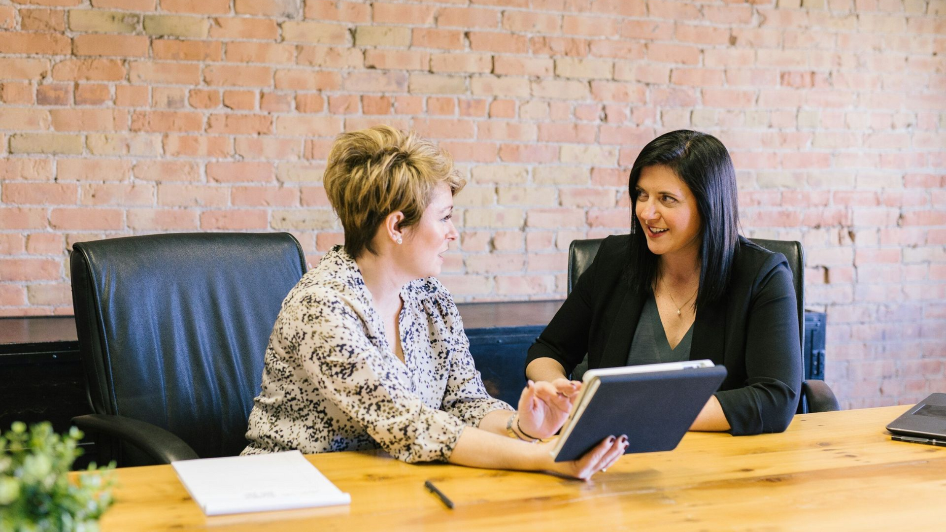 Two women at a wooden table discussing a tablet in front of a brick wall.