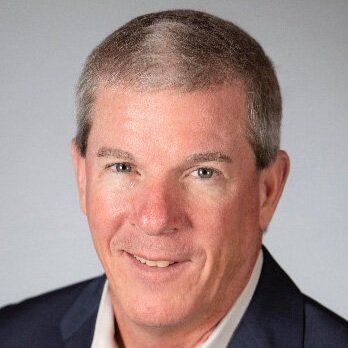 Man with short gray hair, smiling, wearing a dark blue suit jacket, light colored shirt, and a neutral background.