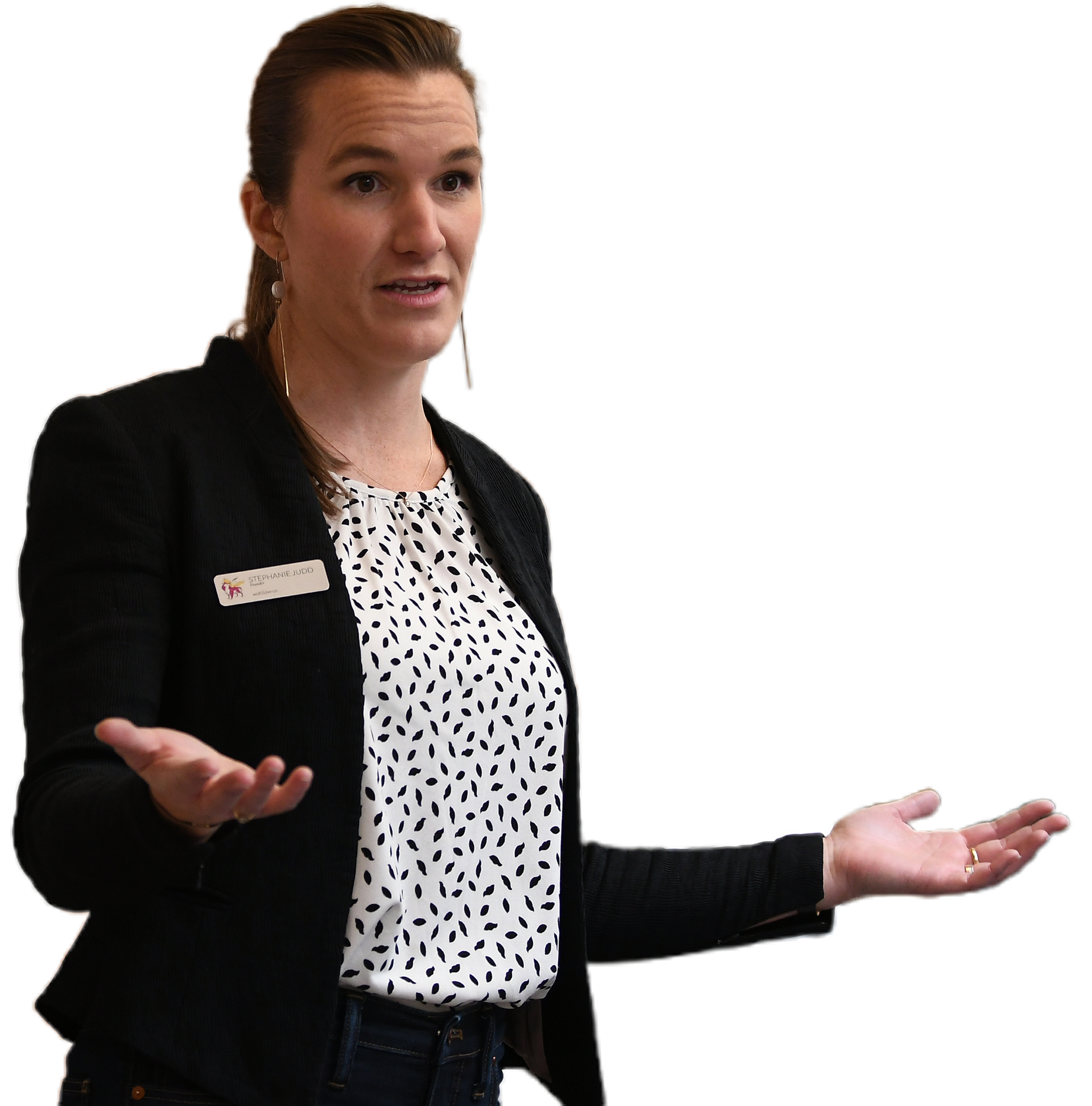 Woman speaking with hands open, wearing black blazer over patterned blouse and jeans.