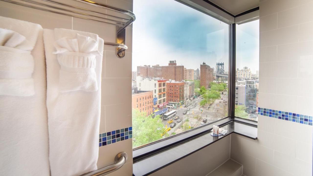 Bathroom with city view, white robes on a rack, buildings, and a bridge visible through the large window.