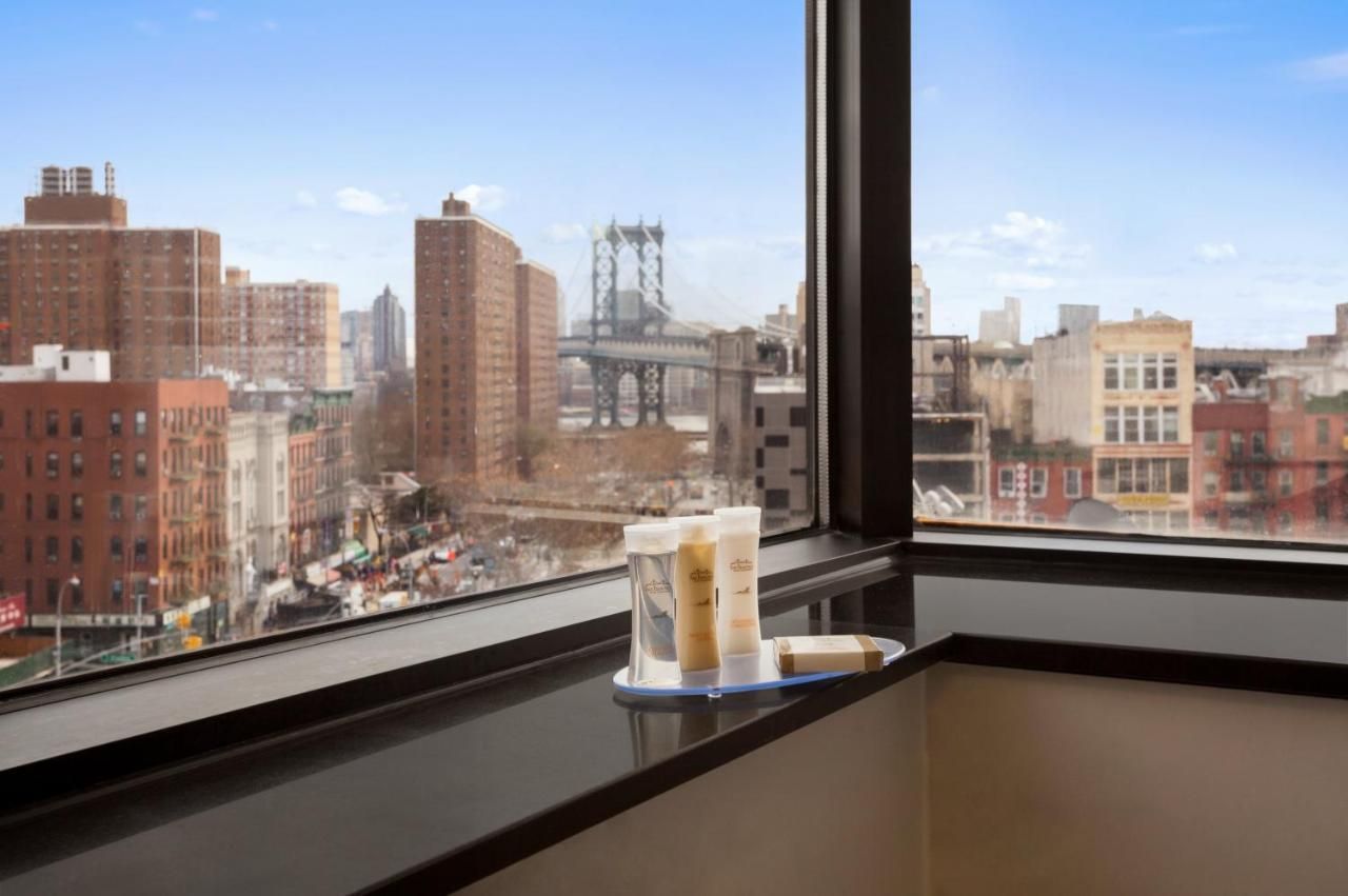City view through a window, toiletries on the sill. The Manhattan Bridge and brick buildings are visible.
