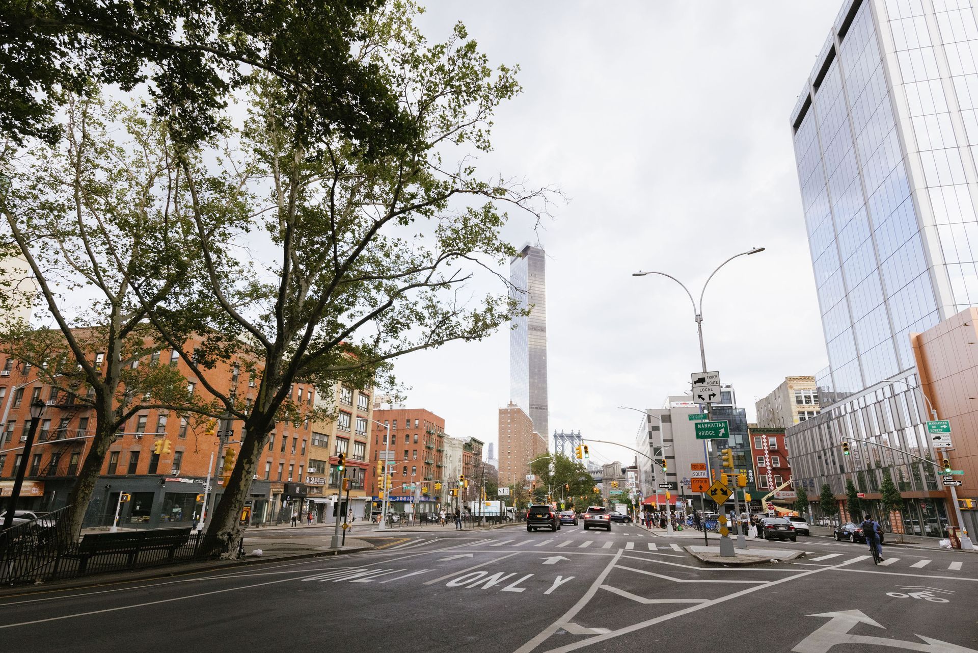 Street scene in NYC with buildings, trees, crosswalk, and vehicles under an overcast sky.