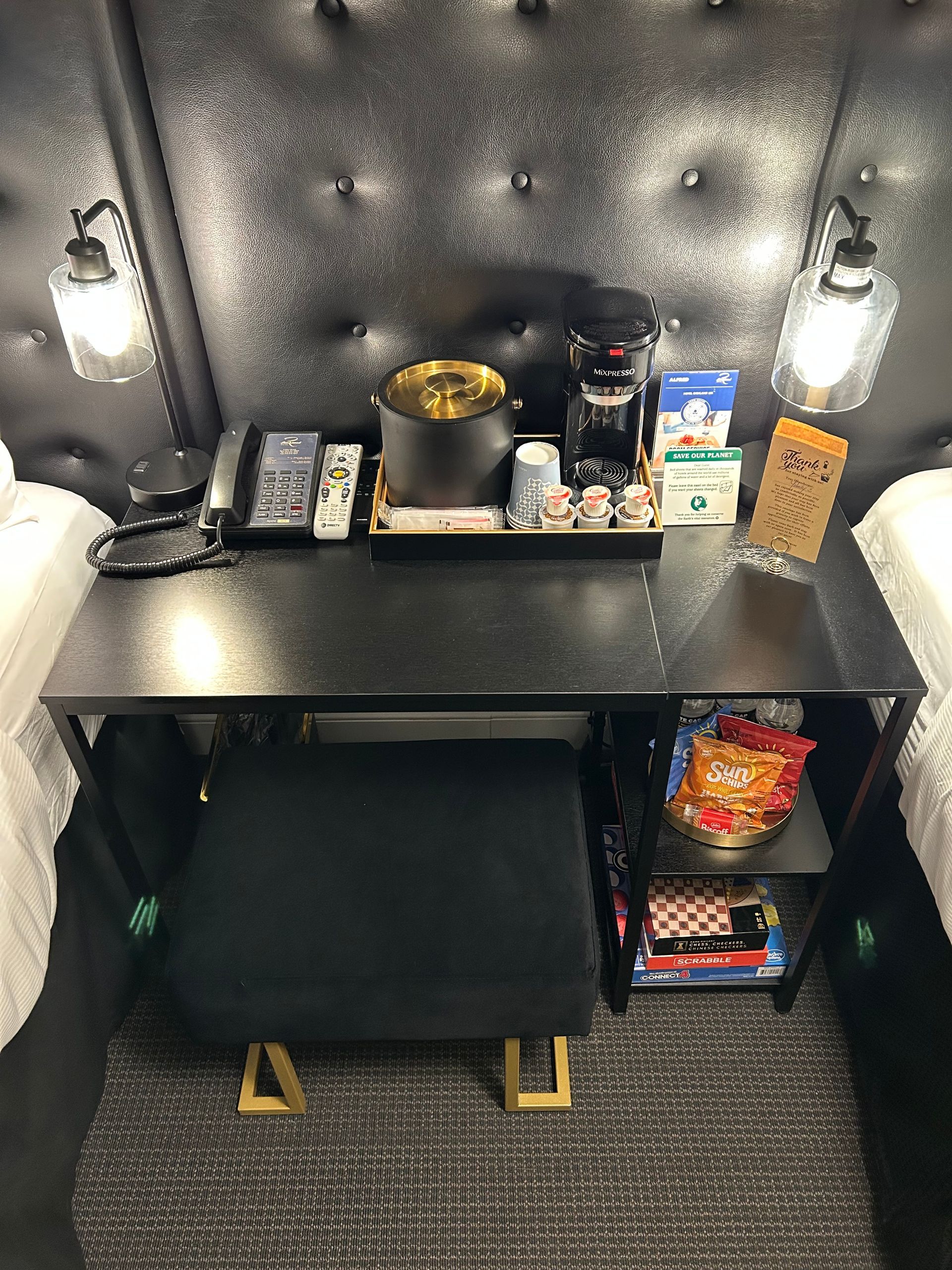 Desk with coffee maker, snacks, and a stool between two beds, under a buttoned headboard.