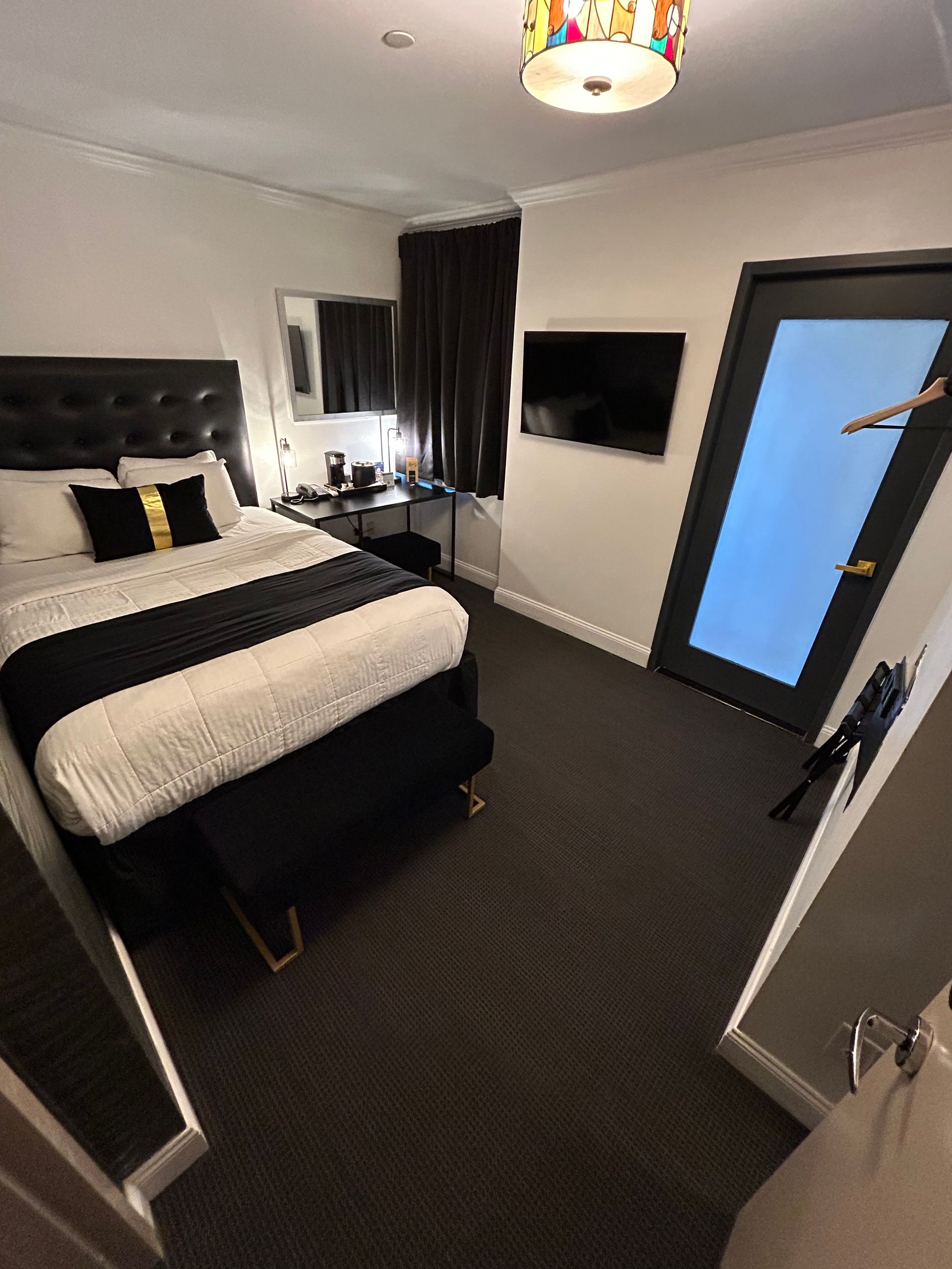 Hotel room with a black and white bed, TV, and open door. Dark carpet and walls, colorful light fixture.