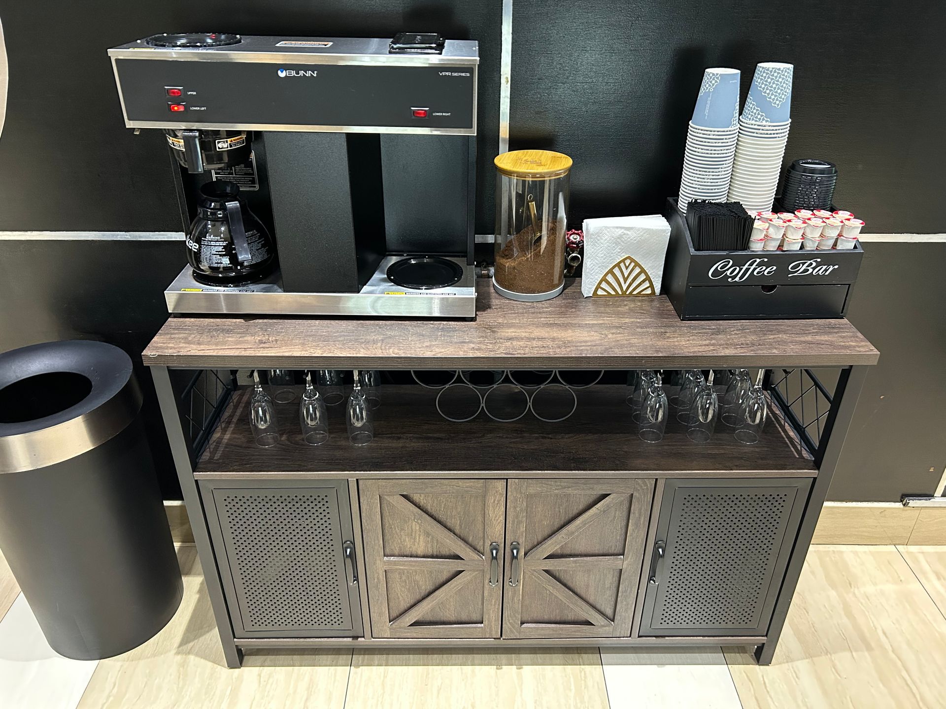 Coffee station: coffee maker on a rustic cabinet with coffee supplies, glasses, and a trash can.