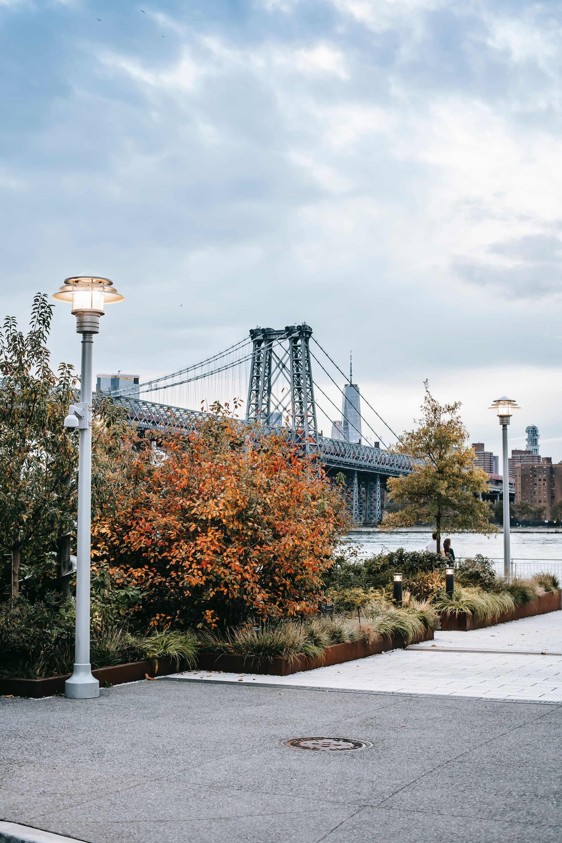 Park with autumn foliage, lit streetlamps, and the Williamsburg Bridge in the background on a cloudy day.