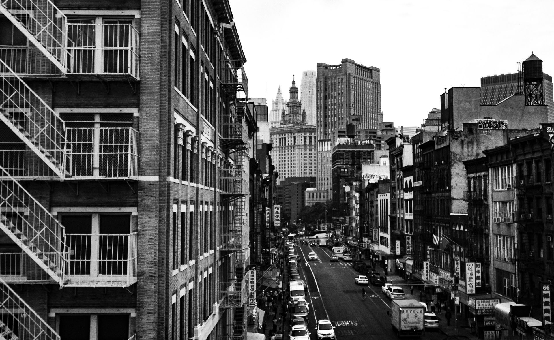 Black and white urban street scene. Tall brick buildings line the road. Cars and trucks travel.