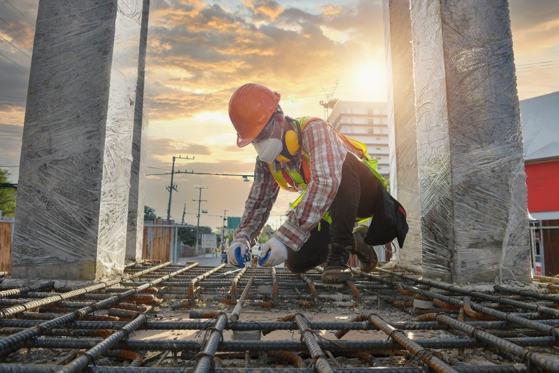 Construction worker in a hard hat and safety gear tying rebar, with the sun setting behind concrete pillars.