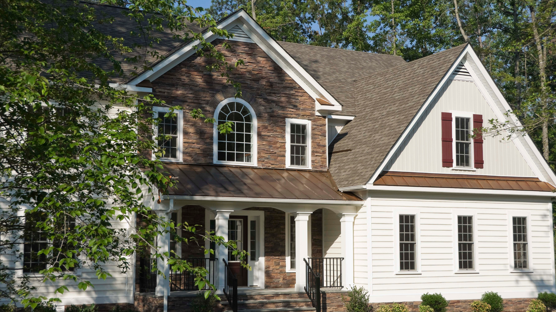Two-story house with brick and white siding. Brown roof and red shutters, front porch, trees in background.
