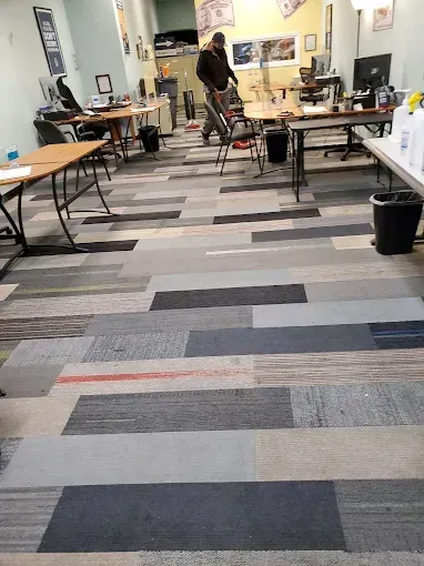 Person vacuuming carpet tiles in an office. Tables, computers, and supplies visible.