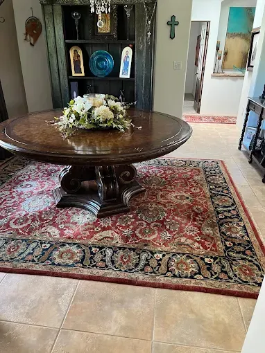 A round wooden table with floral arrangement on a patterned rug in an entryway with a shelf.