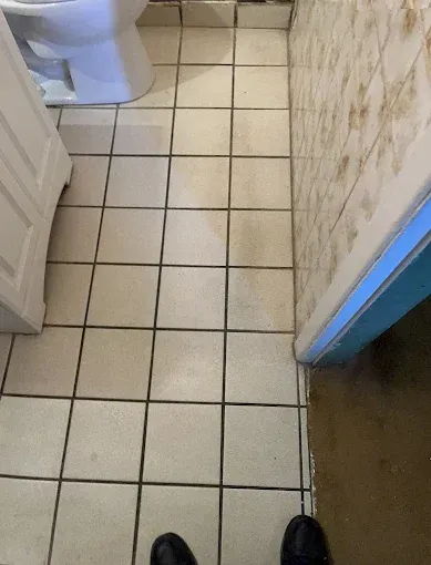 Tiled bathroom floor with a toilet, doorframe, and a person's feet visible. The floor is beige.