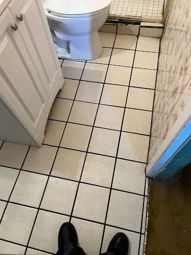 Bathroom floor with square, off-white tiles. Toilet and vanity visible. Black shoes in the foreground.