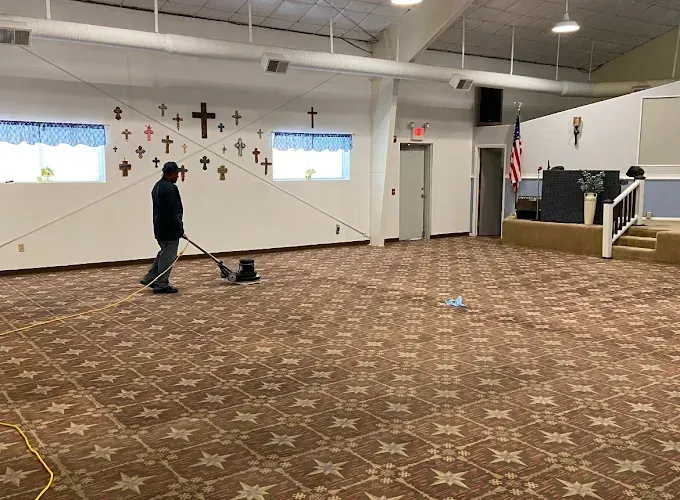 Person using a carpet cleaner in a church hall with crosses on the wall and a small stage.