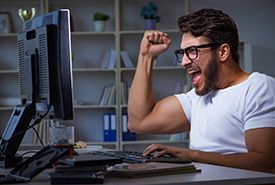 Man with glasses clenches fist, celebrating at a computer, white shirt, office setting.