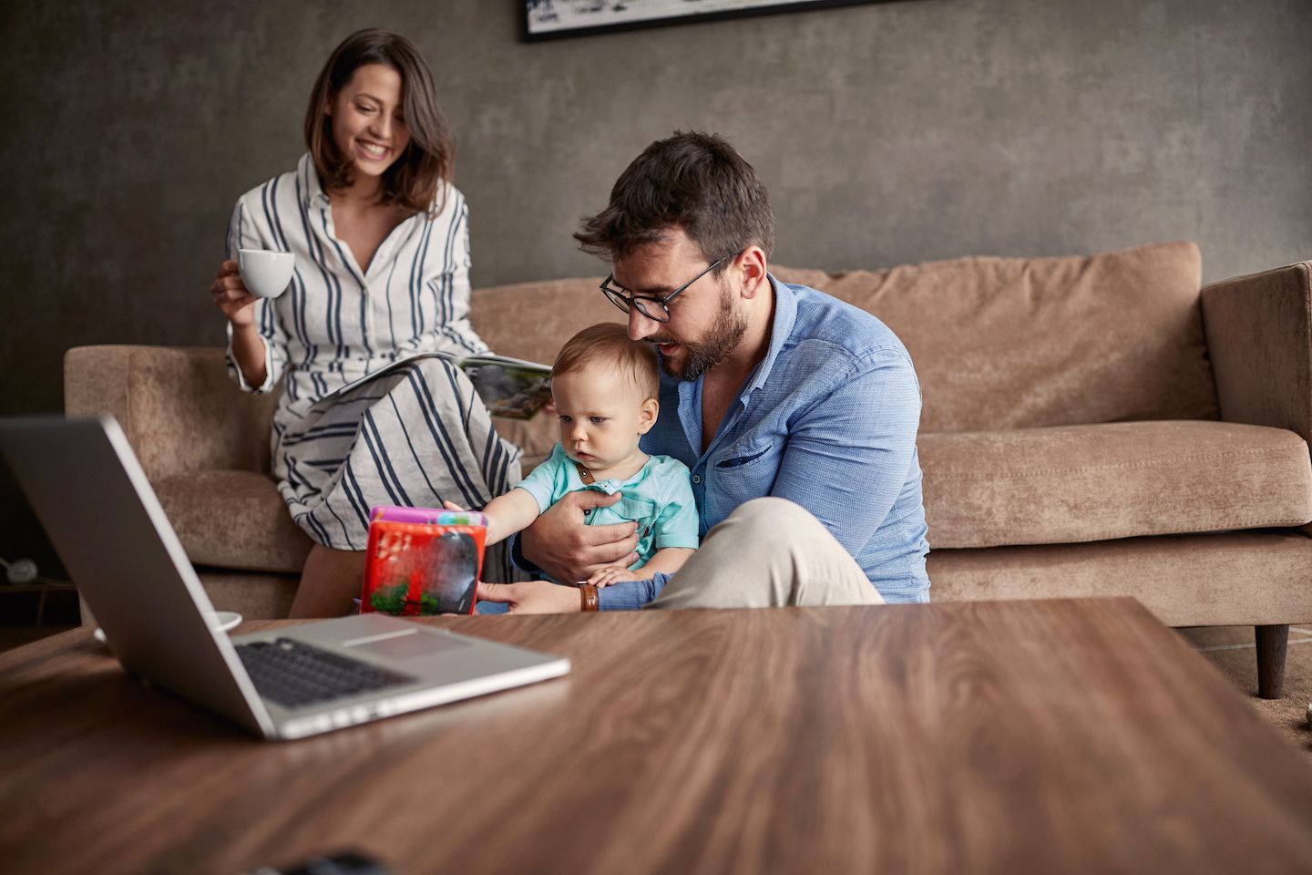 A family sits in a living room: mother with a coffee, father with a baby, laptop on table.