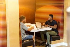 Two people sit at a small table inside a booth, working. One uses a laptop. The walls are orange.
