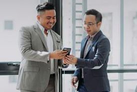 Two men in suits looking at a phone, smiling indoors near a window.