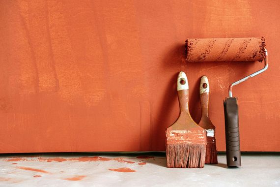 A paint roller and two brushes are sitting in front of an orange wall.