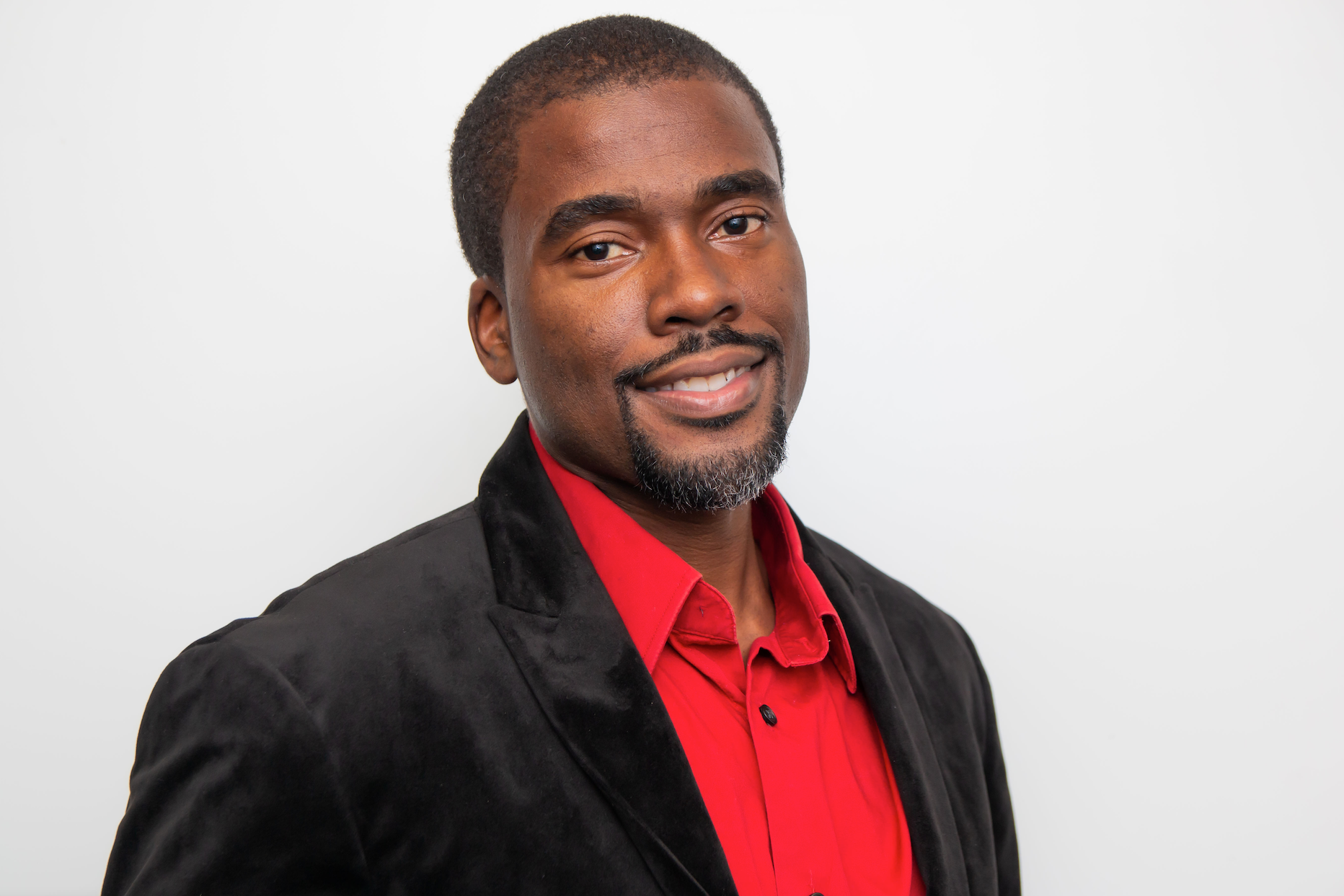 A man wearing a red shirt and a black jacket is smiling for the camera.