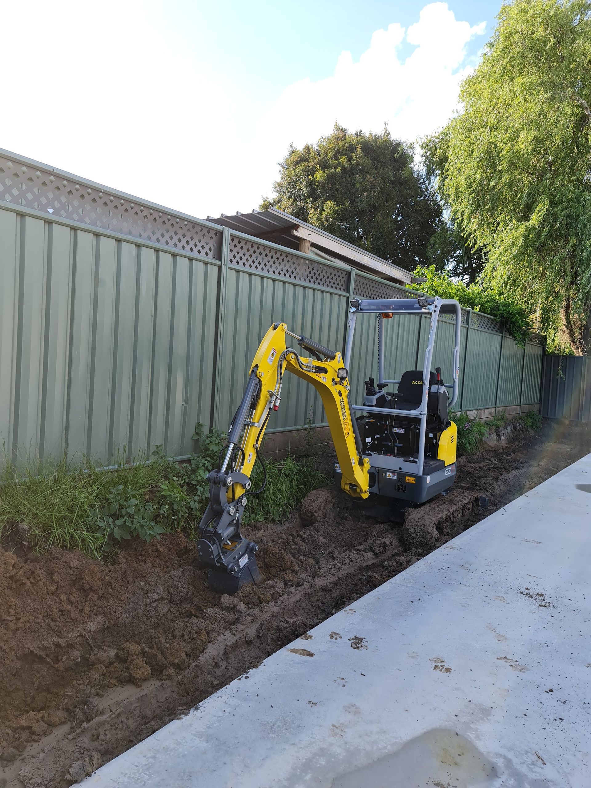 A small yellow excavator is digging a hole in the dirt next to a fence.