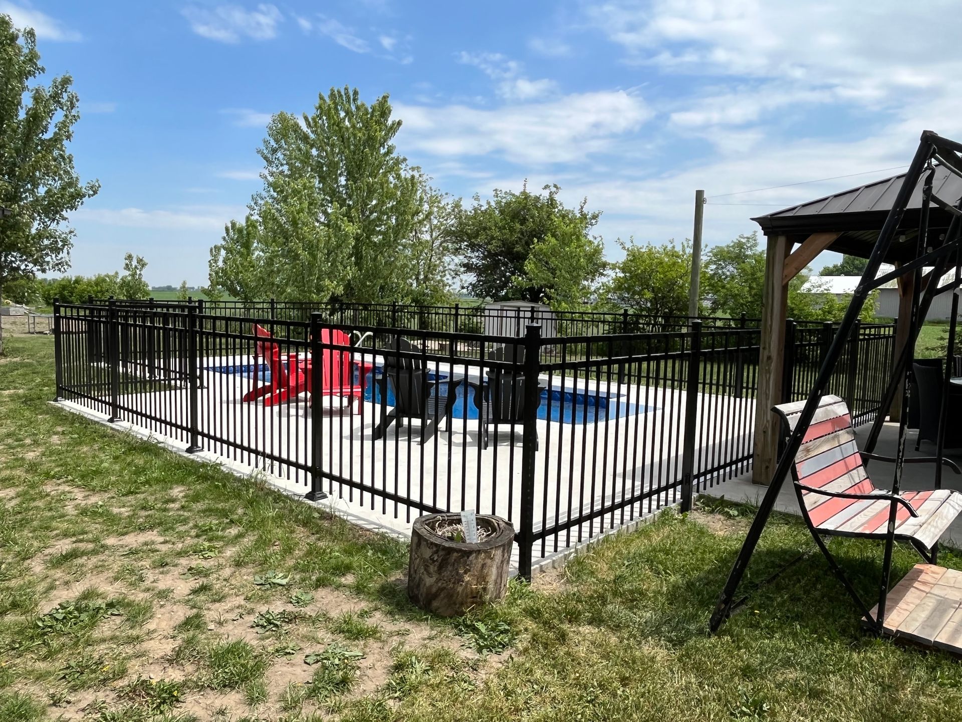 Une clôture noire entoure une piscine avec des chaises et un gazebo.