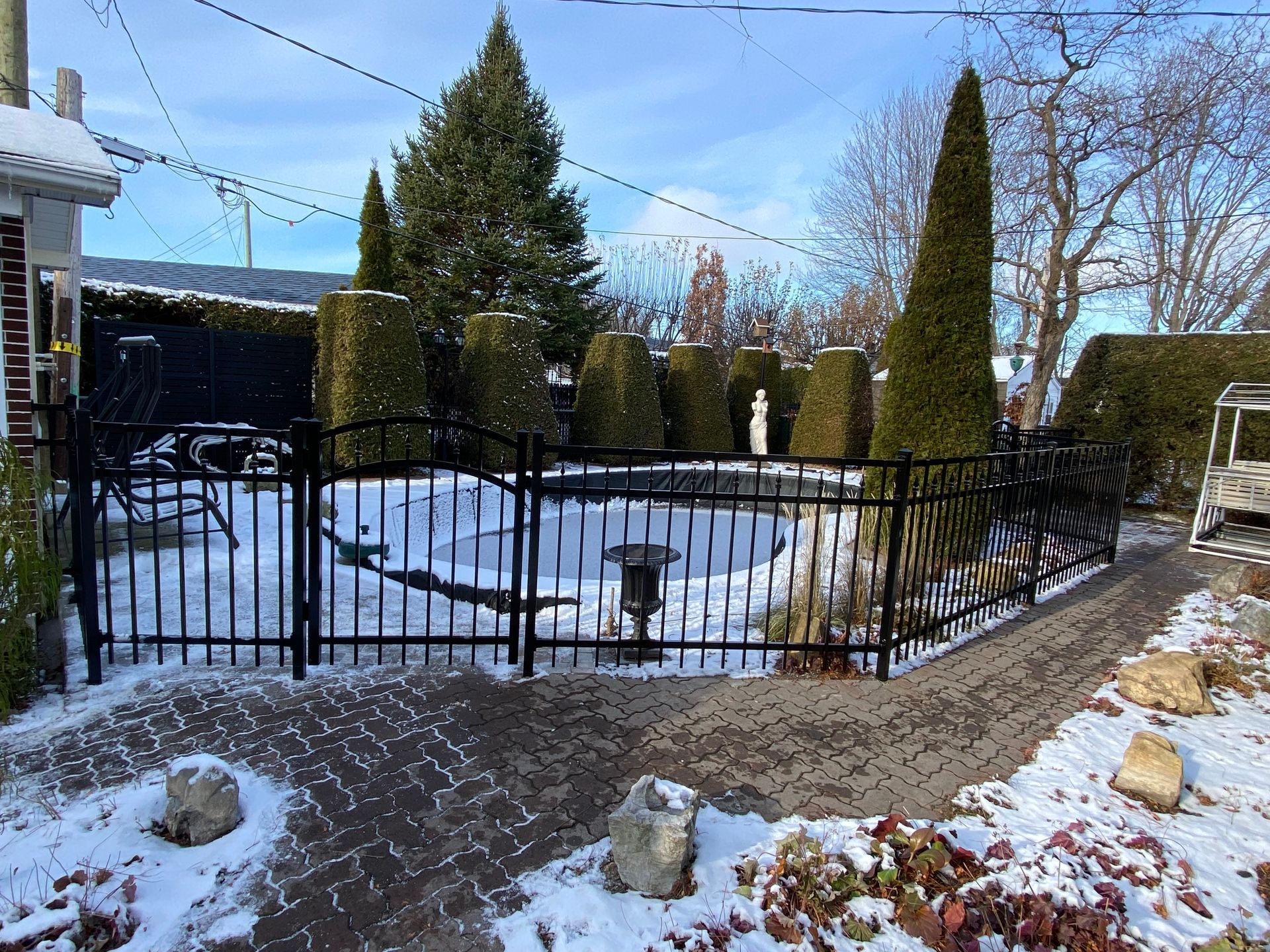 Une clôture noire entoure une cour enneigée avec des arbres et des buissons.