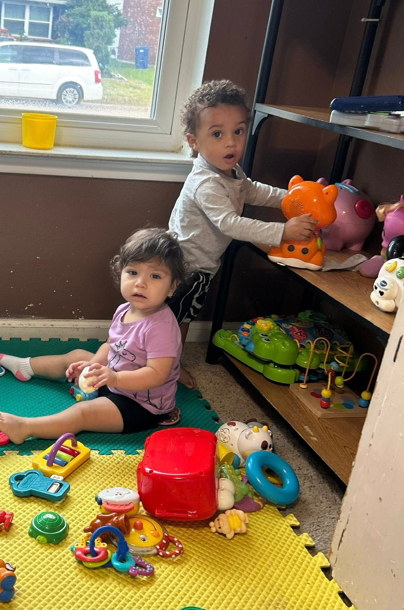 Two young children playing with toys in a room. One sits on the floor with a toy, the other holds a toy on a shelf.