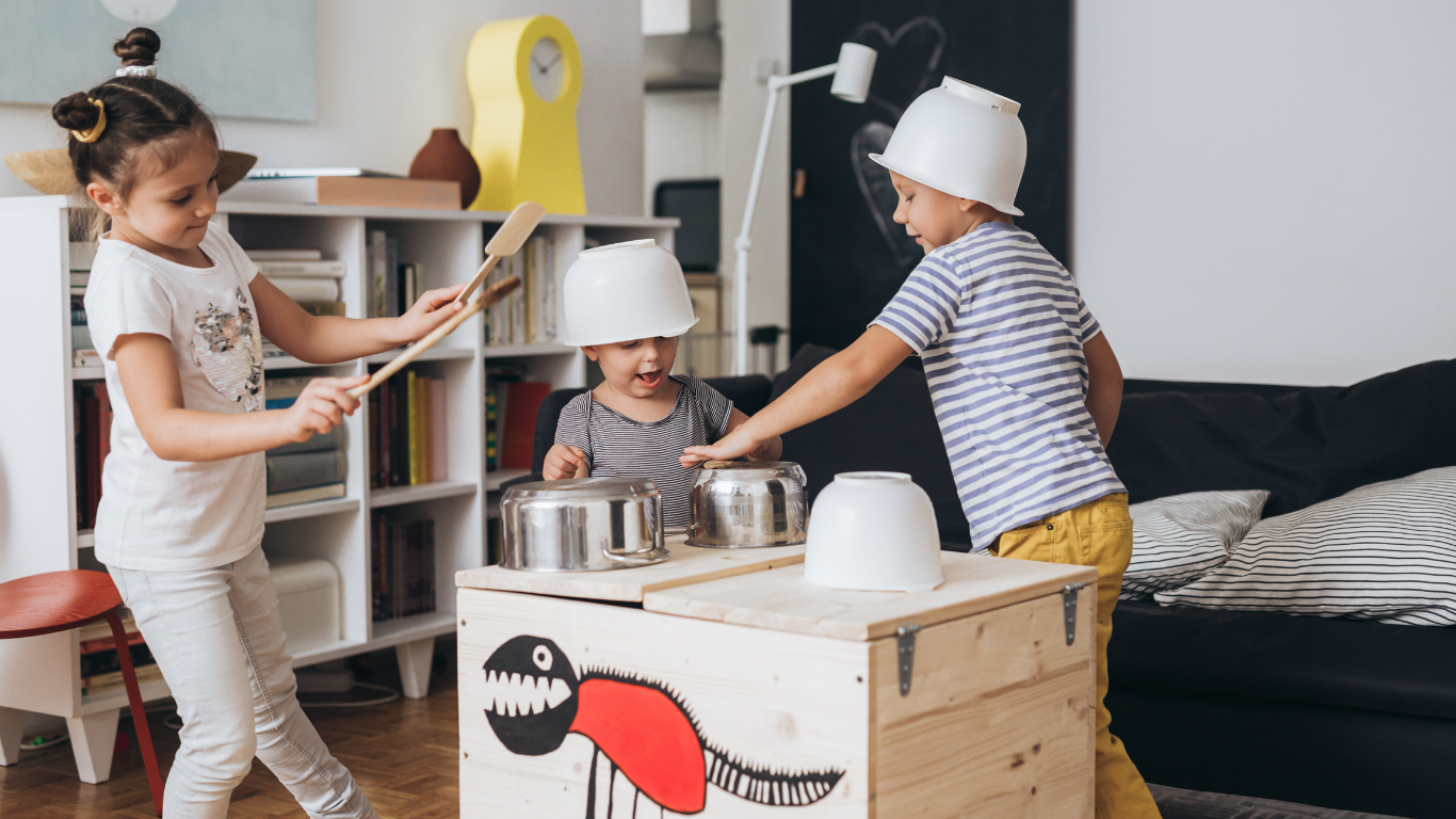 Three children playing with pots and pans on a wooden box, wearing helmets, in a living room.