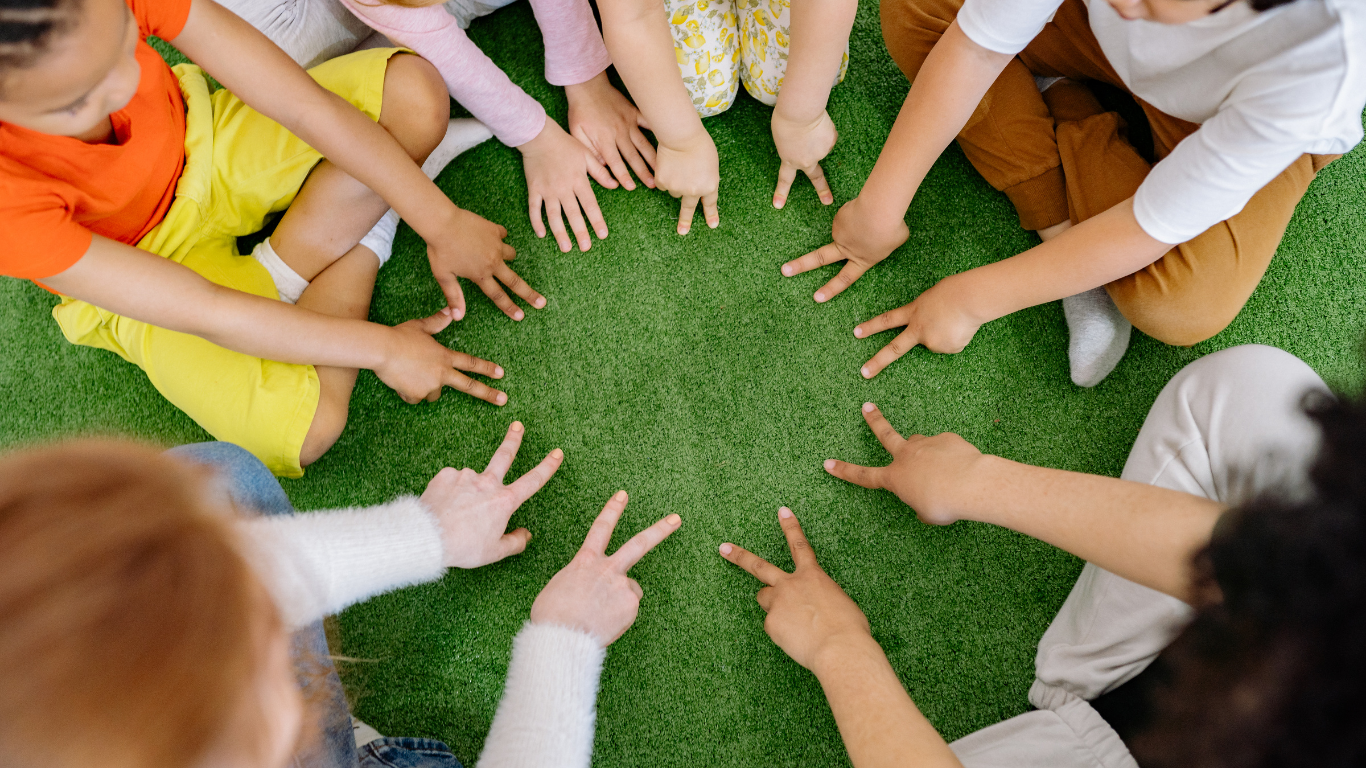 Children's hands in a circle, showing "peace" signs. They are on a green surface.