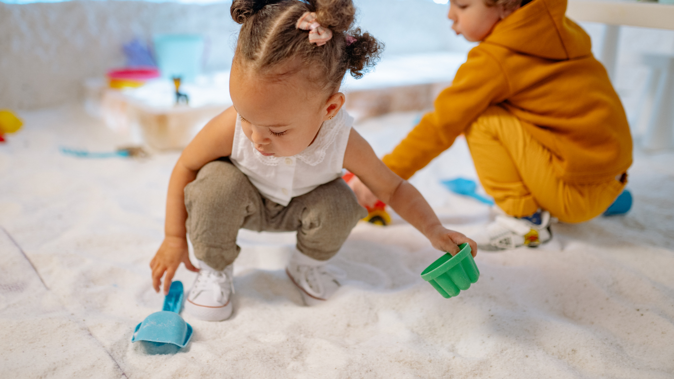 Two toddlers playing with toys in a white salt room. One crouches, reaching for toys. The other sits, also reaching.
