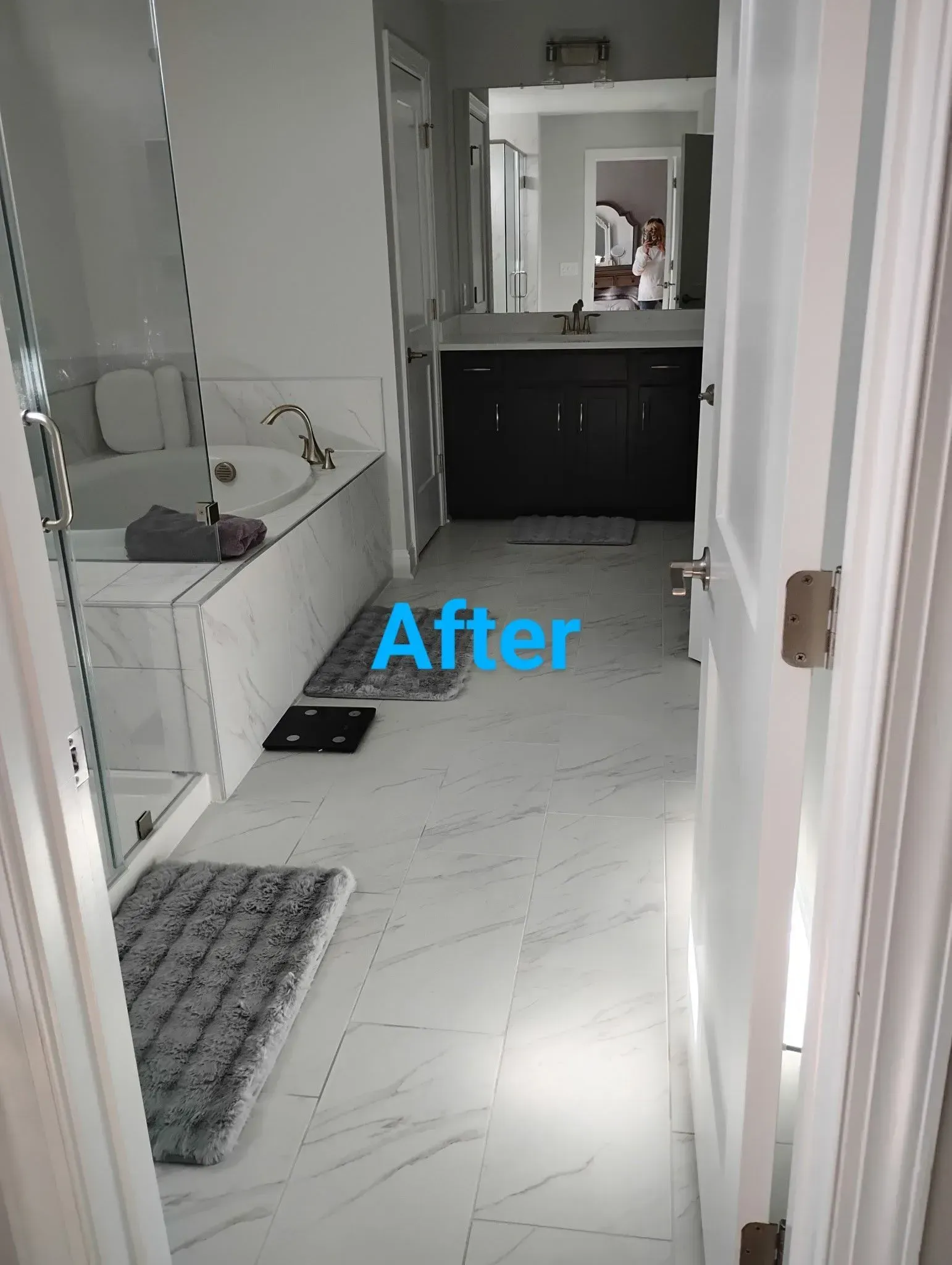 White marble-floored bathroom with bathtub, shower, dark vanity, and grey rugs. Doorway on right.
