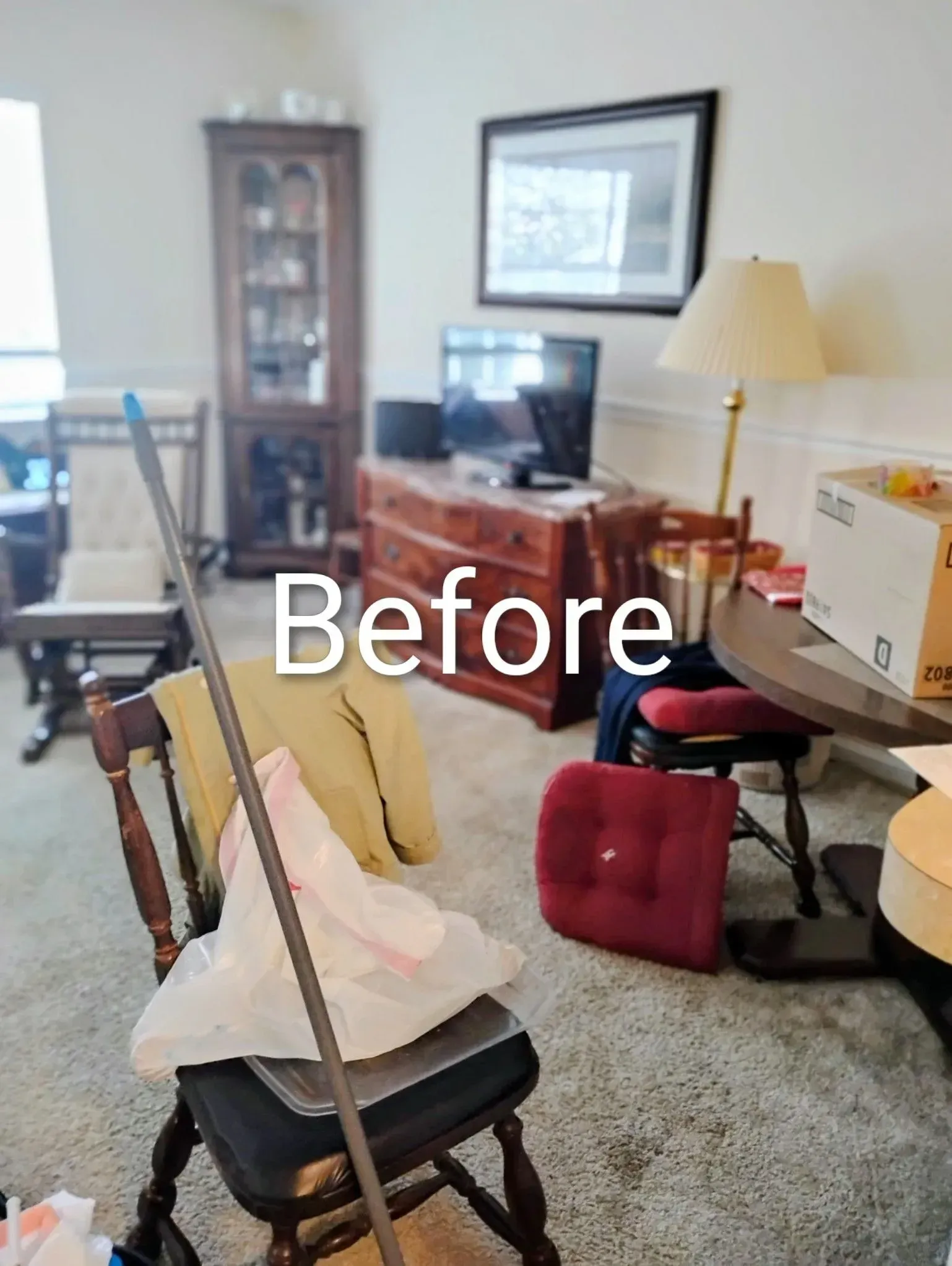 Cluttered living room. Chair holding items, cabinet, TV, lamp, table, chairs, beige carpet, and white walls.